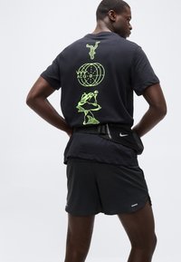 Άνδρας ντυμένος με μαύρο αθλητικό σύνολο με νέον πράσινα αφηρημένα γραφικά στην πλάτη του μπλουζάκι, στέκεται με τα χέρια στη μέση.