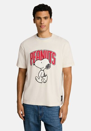 Junger Mann trägt ein weißes T-Shirt mit einem roten "PEANUTS" Logo und einer Snoopy-Figurengrafik, kombiniert mit blauen Jeans, steht vor weißem Hintergrund.