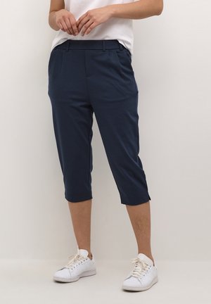 Personne portant un pantalon court bleu marine, des baskets blanches et un haut blanc à manches courtes, debout devant un fond uni.