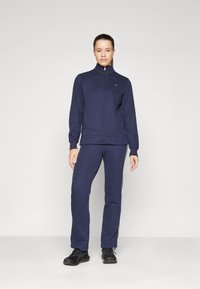 Jūrų laivyno zip-up megztinis ir derančios kelnės iš minkštos medžiagos, su liesu kirpimu. Yra aukšta apykaklė, elastingi rankogaliai ir šoninės juostelės. Juodi batai.