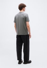 Άντρας με σγουρά μαλλιά στέκεται με την πλάτη γυρισμένη, φοράει γκρι κοντομάνικο πουκάμισο, φαρδιά μαύρα παντελόνια και μαύρα παπούτσια μπροστά σε απλό φόντο.