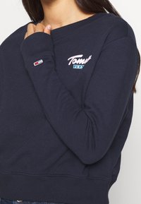 Mörkblå sweatshirt i mjukt tyg, med rundad halsringning och ribbade ärmslut. Inkluderar broderad "Tommy Jeans" logo i rosa och blått.