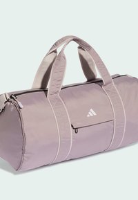 Bolsa de viaje de tela lavanda clara con dos asas superiores, cierre de cremallera, bolsillo frontal y detalle de logo de marca en blanco. Textura suave.