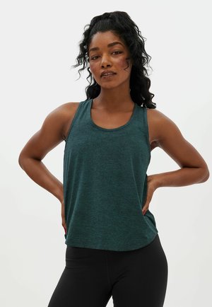 Junge Frau mit lockigem dunklem Haar, die ein ärmelloses, türkisfarbenes Oberteil und schwarze Leggings trägt, mit Händen in der Hüfte vor einem schlichten Hintergrund steht.