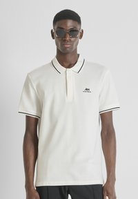 Chemise polo blanche avec des accents noirs au col et aux manches, arborant un petit logo noir sur la poitrine, fabriquée en tissu texturé.