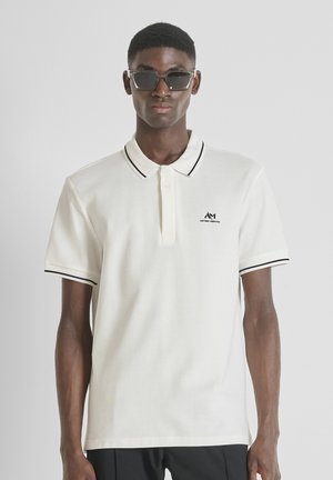 Camisa polo blanca con detalles en negro en el cuello y las mangas, con un pequeño logo negro en el pecho, hecha de una tela texturizada.