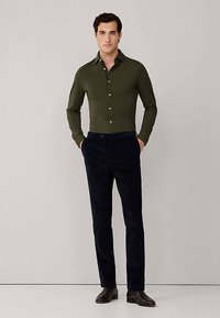 Mørkegrøn skjorte med lange ærmer og knapper foran, kombineret med mørkeblå fløjlsbukser. Modellen bærer brune lædersko. Simpelt design.