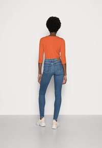 Top naranja de manga larga con cuello redondo, combinado con jeans ajustados de talle alto en azul. Presenta dos bolsillos traseros y una etiqueta de cuero en la cinturilla.