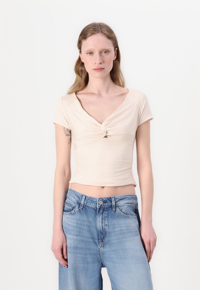 Top beige aderente con maniche corte, scollo a V e dettaglio frontale attorcigliato, abbinato a jeans a gamba larga blu chiaro. Tessuto liscio e elastico.