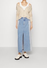 Beiger Cardigan mit Knöpfen, getragen über einem weißen Tanktop. Hellblaue Jeans-Maxi-Rock mit vorderem Schlitz und kontrastierenden Seitenteilen. Cremefarbene Schuhe.