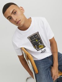 Camiseta blanca de algodón con un gráfico en negro y amarillo de un esqueleto tocando la guitarra junto a una ventana. Modelo sentado en una silla de metal.