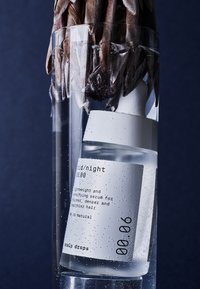 Bouteille en verre transparent avec étiquette blanche immergée dans l'eau, présentant une forme arrondie, un design minimaliste et des bulles visibles. Texte : "gouttes pour cuir chevelu."