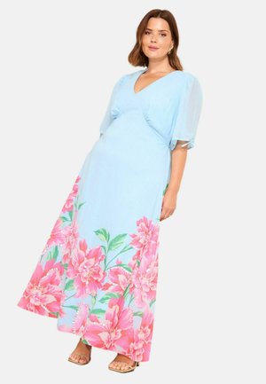 Frau trägt ein hellblaues Maxi-Kleid mit V-Ausschnitt, transparenten Puffärmeln und großem pinkfarbenem Blumenmuster entlang des Saums, steht vor weißem Hintergrund.