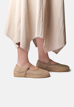 Fødder iført beige vævede Mary Jane-flade sko med ankelremme, kombineret med et flydende lysefarvet skørt med ujævn kant.