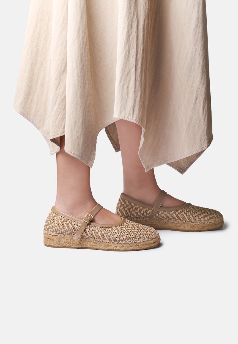 Pieds portant des ballerines Mary Jane tissées beige avec des brides à la cheville, assorties à une jupe fluide de couleur claire avec un ourlet irrégulier.