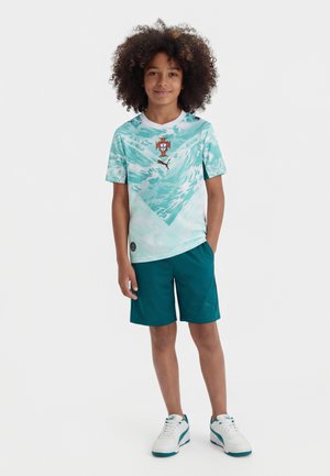 Αγόρι με σγουρά μαλλιά που φοράει μπλε-τιρκουάζ και λευκή φανέλα ποδοσφαίρου με σχέδιο, τυρκουάζ σορτς και λευκά αθλητικά παπούτσια, στέκεται με το ένα χέρι στην τσέπη.