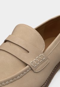 Mocassin en daim beige avec bride décorative et couture blanche le long du bord, semelle marron visible en dessous.