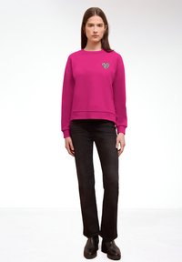 Jeune femme debout, portant un sweat-shirt rose vif avec un détail en forme de cœur imprimé léopard, un jean noir et des bottes noires sur un fond blanc.