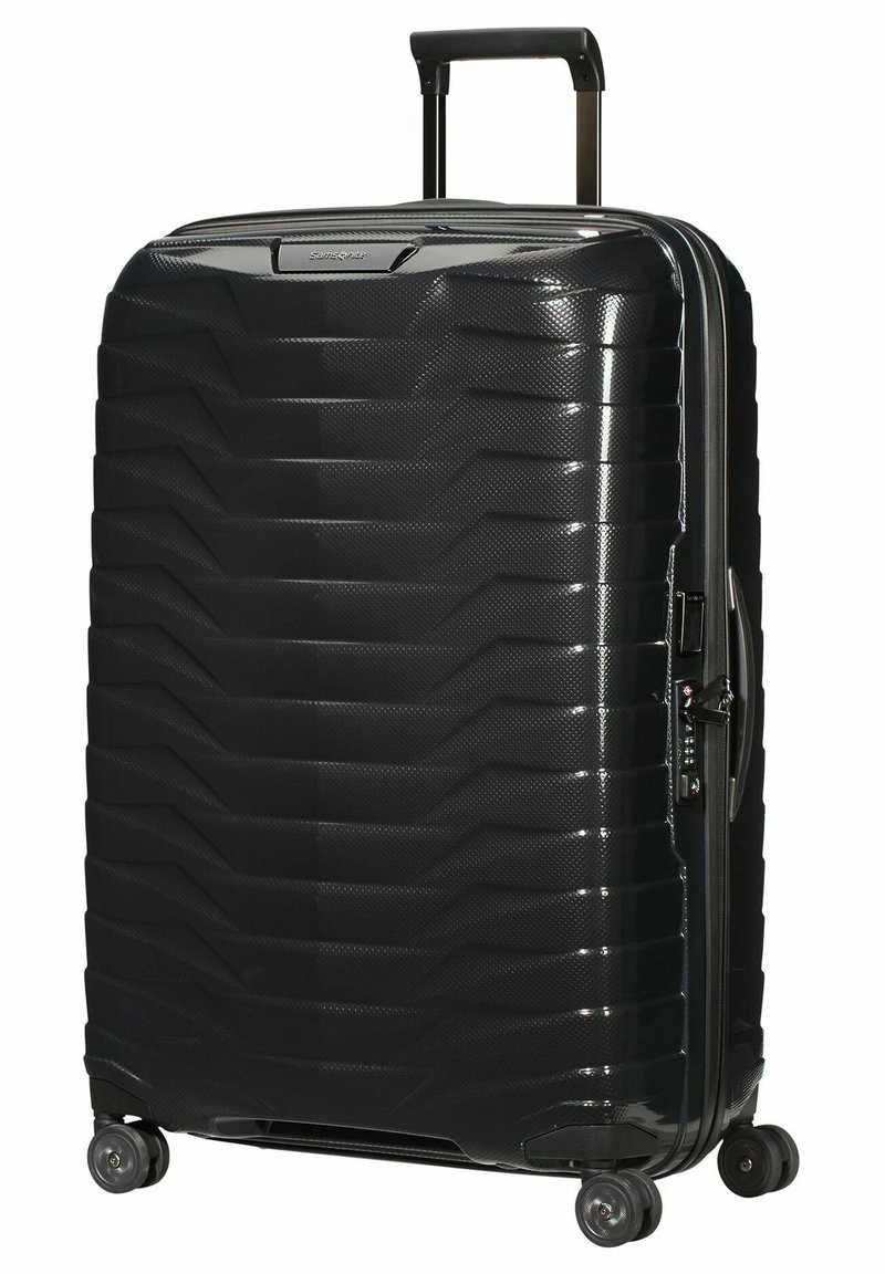Valise rigide noire avec une surface texturée, motif géométrique, poignée extensible, quatre roues pivotantes et ouverture zippée.