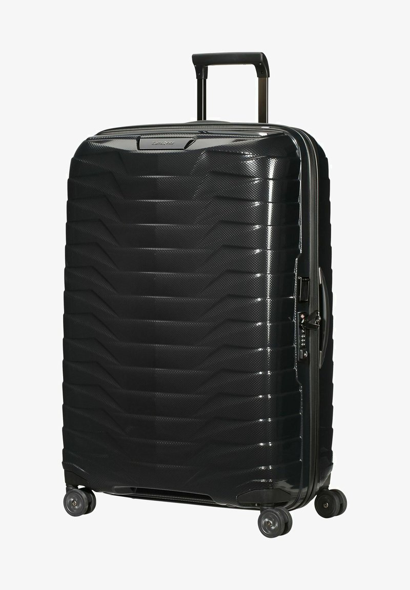 Valise rigide noire avec une surface texturée, motif géométrique, poignée extensible, quatre roues pivotantes et ouverture zippée.