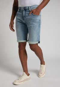 Shorts en denim bleu clair avec des ourlets roulés, cinq poches et des coutures visibles, assortis à des baskets beiges avec une rayure rouge.