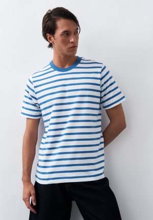 Mand iført hvid T-shirt med korte ærmer og vandrette blå striber, står med den ene hånd bag ryggen og kigger til siden.