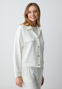 Witte cropped jas van gladde stof, met een gekraagde halslijn, twee borstzakken en gouden knopen.