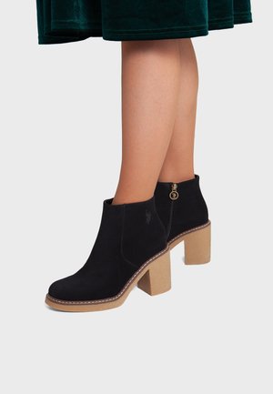Jambes portant des bottines en daim noir avec talons blocs couleur cuir clair et fermeture éclair dorée, associées à une jupe en velours vert foncé.