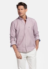 Chemise à rayures rouges et blanches avec une poche, détails bleus contrastants au niveau des poignets, fabriquée en tissu léger. Portée avec un pantalon blanc.
