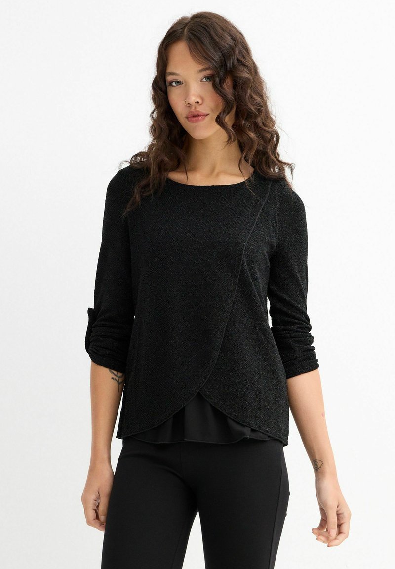 Top negro de manga larga con acabado texturizado, que presenta un dobladillo asimétrico y un diseño en capas. Se lleva sobre una capa inferior plisada negra.