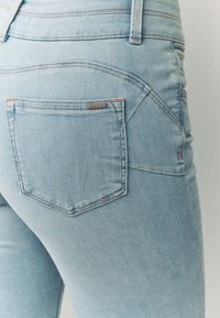 Svijetloplave traper hlače sa glatkom teksturom, standardnim dizajnom džepova i suptilnim naglascima šavova. Pogled s leđa ističe oblikovani struk.