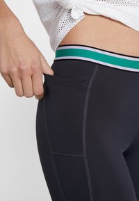 Personne portant des leggings noirs avec une ceinture rayée verte et violette, montrant une poche latérale avec la main à l'intérieur.