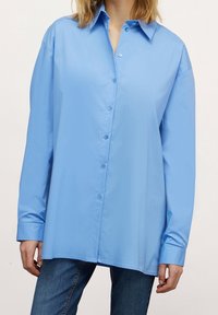 Light blue button-up shirt made of smooth fabric, featuring a pointed collar and long sleeves with cuffs. Model is wearing denim jeans.