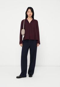 Woman wearing a burgundy long-sleeve top, navy wide-leg pants, black shoes, and carrying a beige shoulder bag, standing against a white background.