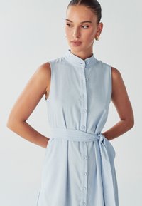 Šviesiai mėlyna, be rankovių suknelė su aukštu apykakle, priekiniais mygtukais ir surištu liemeniu. Pagaminta iš glotnaus audinio su subtilia tekstūra.