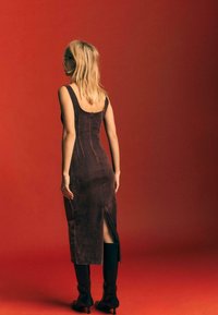 Robe en velours marron avec un dos dégagé et une fente sur le côté, offrant une coupe ajustée qui descend jusqu'au mollet. Le mannequin l'a associée à des bottes noires élégantes.