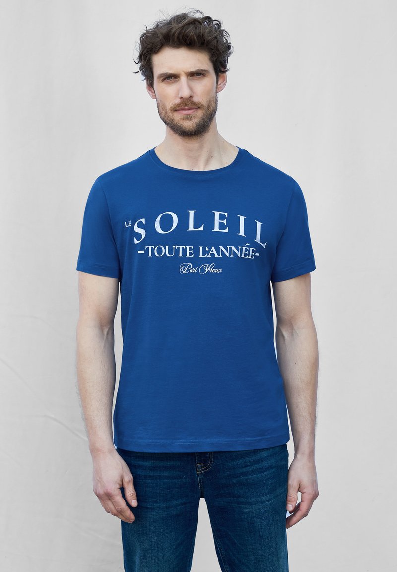 Sininen puuvillapaita lyhyillä hihoilla ja pyöreällä pääntiellä. Edessä keskellä valkoinen tekstimuotoilu "le SOLEIL - toute l'année".