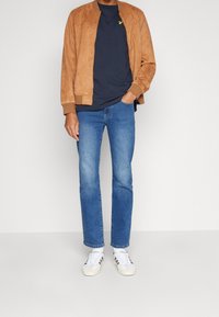 Brązowa bomberka z zamszu, granatowy t-shirt, niebieskie dżinsy, białe trampki; kurtka ma ściągacze przy rękawach i w pasie, t-shirt z małym logo.