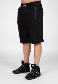 Black athletic shorts with a textured fabric, featuring a red side print and a zippered pocket on the right. Black high-top shoes.