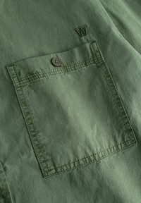 Camicia verde oliva con taschino sul petto, dotata di chiusura a bottoni e dettagli cuciti, con una lettera "W" ricamata sopra la tasca.