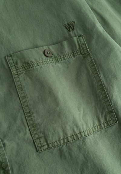 Camicia verde oliva con taschino sul petto, dotata di chiusura a bottoni e dettagli cuciti, con una lettera "W" ricamata sopra la tasca.