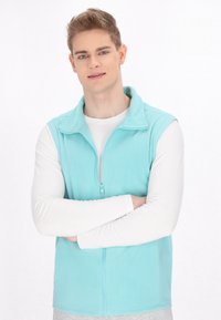 Helles blaufleece-Weste mit hohem Kragen und Frontreißverschluss, getragen über einem weißen Langarmshirt. Weiche Textur, einfaches Design, keine Muster.