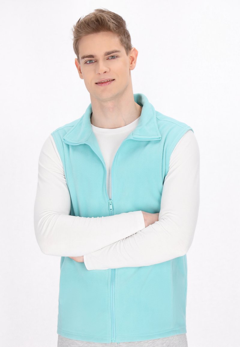 Helles blaufleece-Weste mit hohem Kragen und Frontreißverschluss, getragen über einem weißen Langarmshirt. Weiche Textur, einfaches Design, keine Muster.