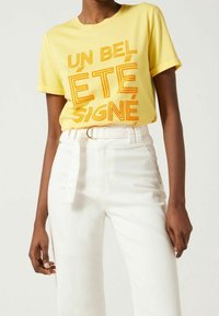 Žluté bavlněné tričko s nápadným oranžovým textem "UN BEL ÉTÉ SIGNÉ." V kombinaci s bílými džínami s vysokým pasem, které zvýrazňují pás.