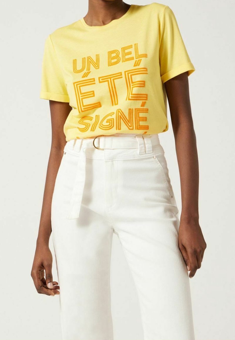 Žluté bavlněné tričko s nápadným oranžovým textem "UN BEL ÉTÉ SIGNÉ." V kombinaci s bílými džínami s vysokým pasem, které zvýrazňují pás.