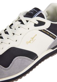 White, black, and grey sneaker with contrasting textures. Features lace closure and a golden logo on the side. Rubber sole, athletic design.