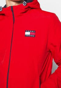 Rød hættejakke lavet af blødt stof, med lynlåslukning og et rektangulært mærke med "TOMMY JEANS"-logo i marineblå og hvide nuancer.