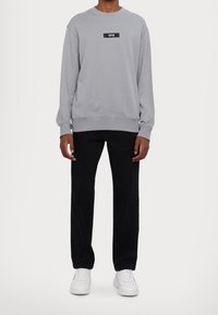 Grå sweatshirt med en svart logotyp på bröstet, tillsammans med svarta raka byxor och vita sneakers. Sklämt tyggtextur.