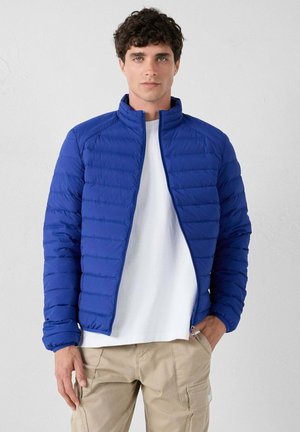 Un giovane che indossa una giacca trapuntata blu sopra una camicia bianca e pantaloni cargo beige, in piedi di fronte a uno sfondo grigio chiaro uniforme.