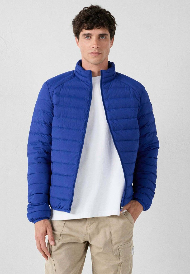 Jonge man in een blauwe gewatteerde jas over een wit shirt en beige cargo broek, staand tegen een effen lichtgrijze achtergrond.